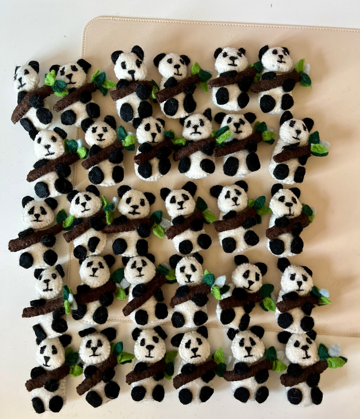 Rows of small felt pandas with black and white markings holding brown branches adorned with green leaves on a beige surface