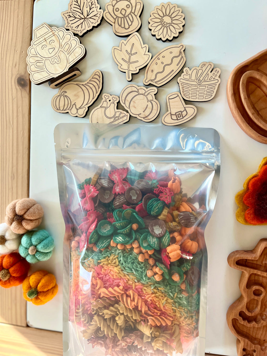 Clear resealable bag filled with multicolored pasta and fall-themed shapes surrounded by wooden autumn tokens and felt pumpkins