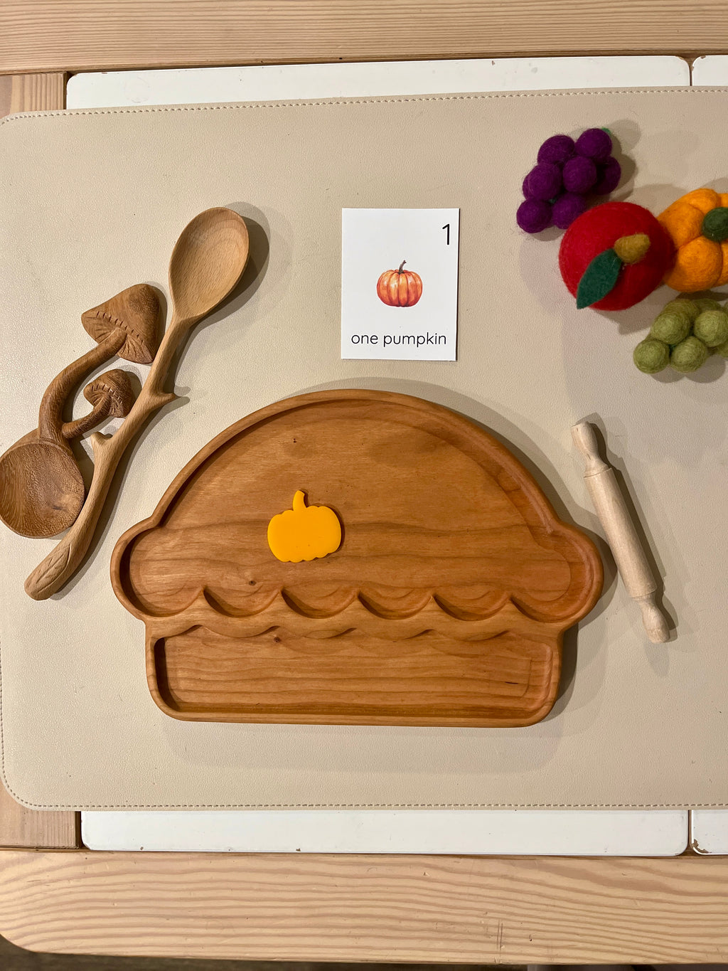Wooden pie-shaped tray with a small yellow felt pumpkin, mushroom-shaped spoons, rolling pin, and felt fruit on beige surface