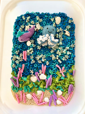 Sensory tray with blue beads, felt octopus and whale, pasta sea creatures, shells, green moss, and colorful clay seaweed pieces.