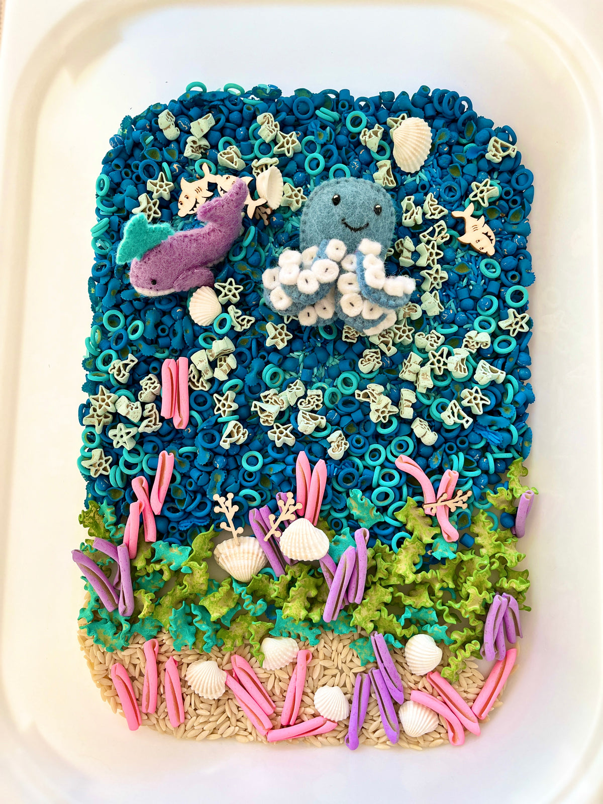 Sensory tray with blue beads, felt octopus and whale, pasta sea creatures, shells, green moss, and colorful clay seaweed pieces.