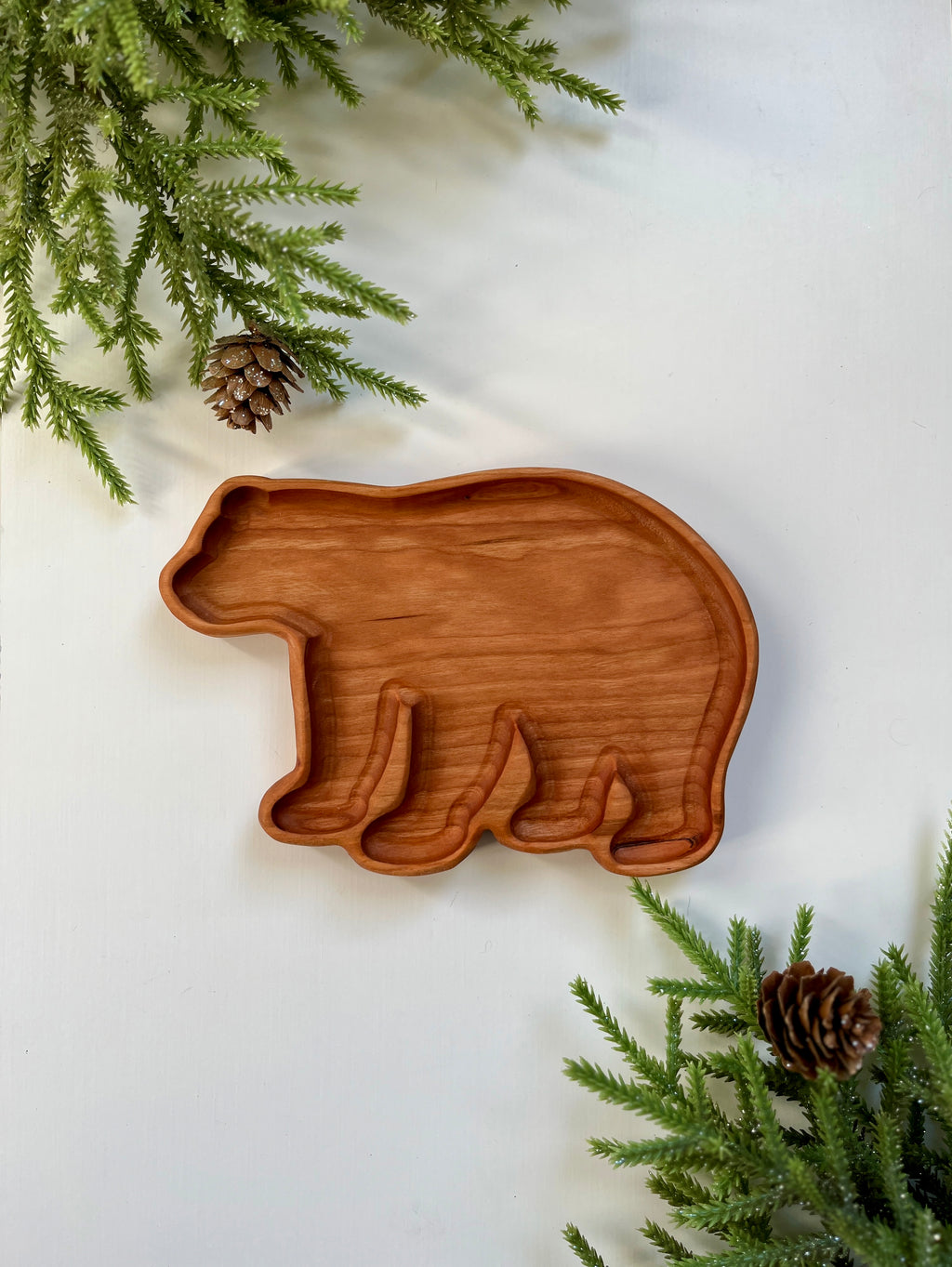 Wooden tray carved in the shape of a bear surrounded by pine branches and pine cones on white surface