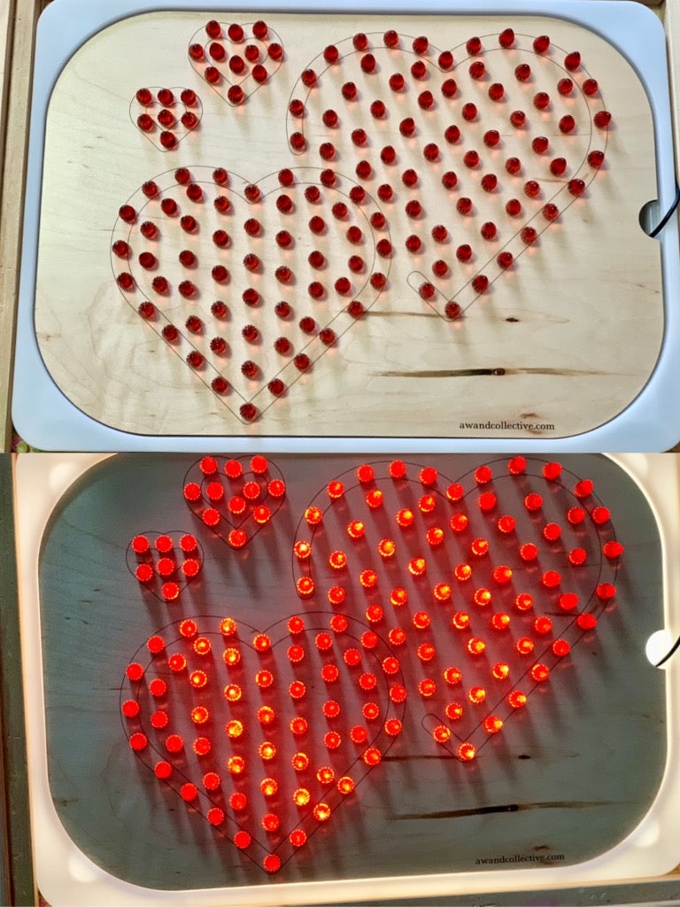Wooden board with four heart outlines filled with removable red pins, shown unlit and glowing in a light box.