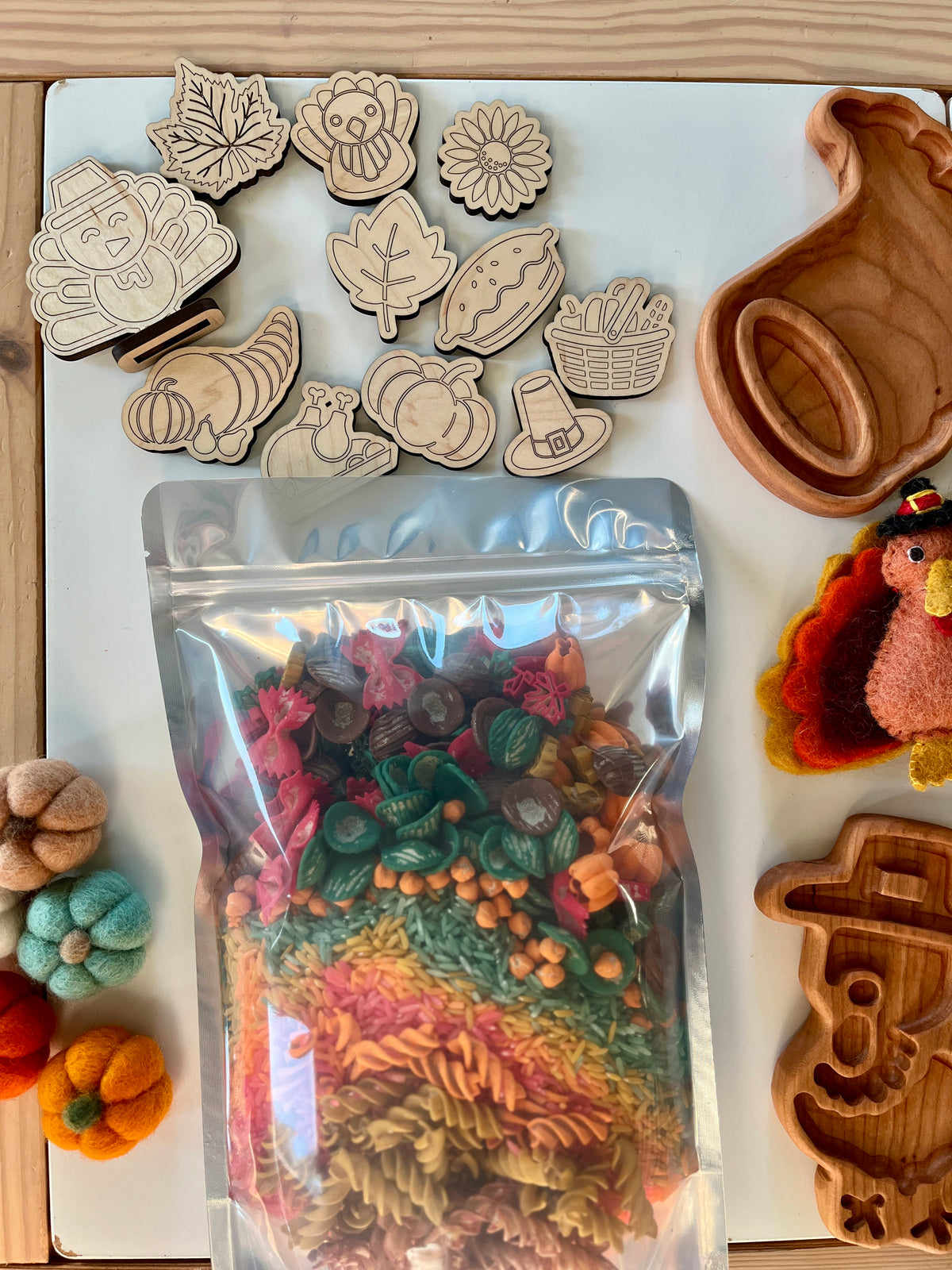 Clear bag with colorful fall-themed pasta shapes surrounded by wooden tokens and autumn craft items on white surface