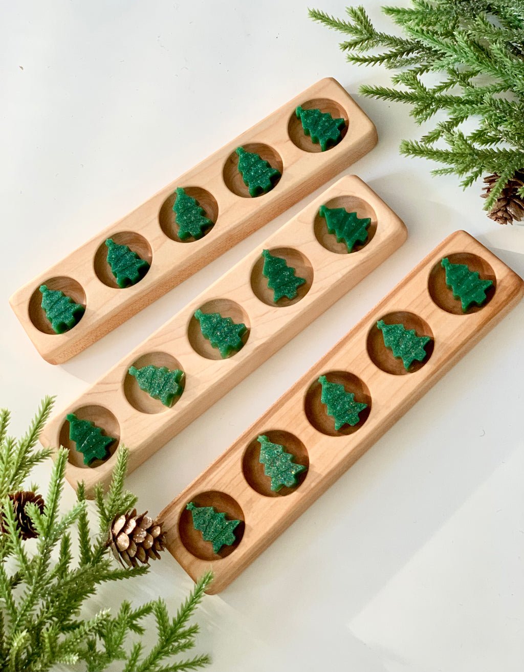 Three wooden five frame sensory trays with green tree-shaped counting gems on white surface with pine branches and pinecones