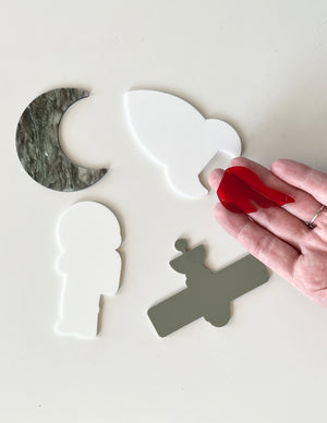 Hand holding red acrylic flame piece above white astronaut, gray satellite, white bird, and green marble crescent moon shapes on white surface