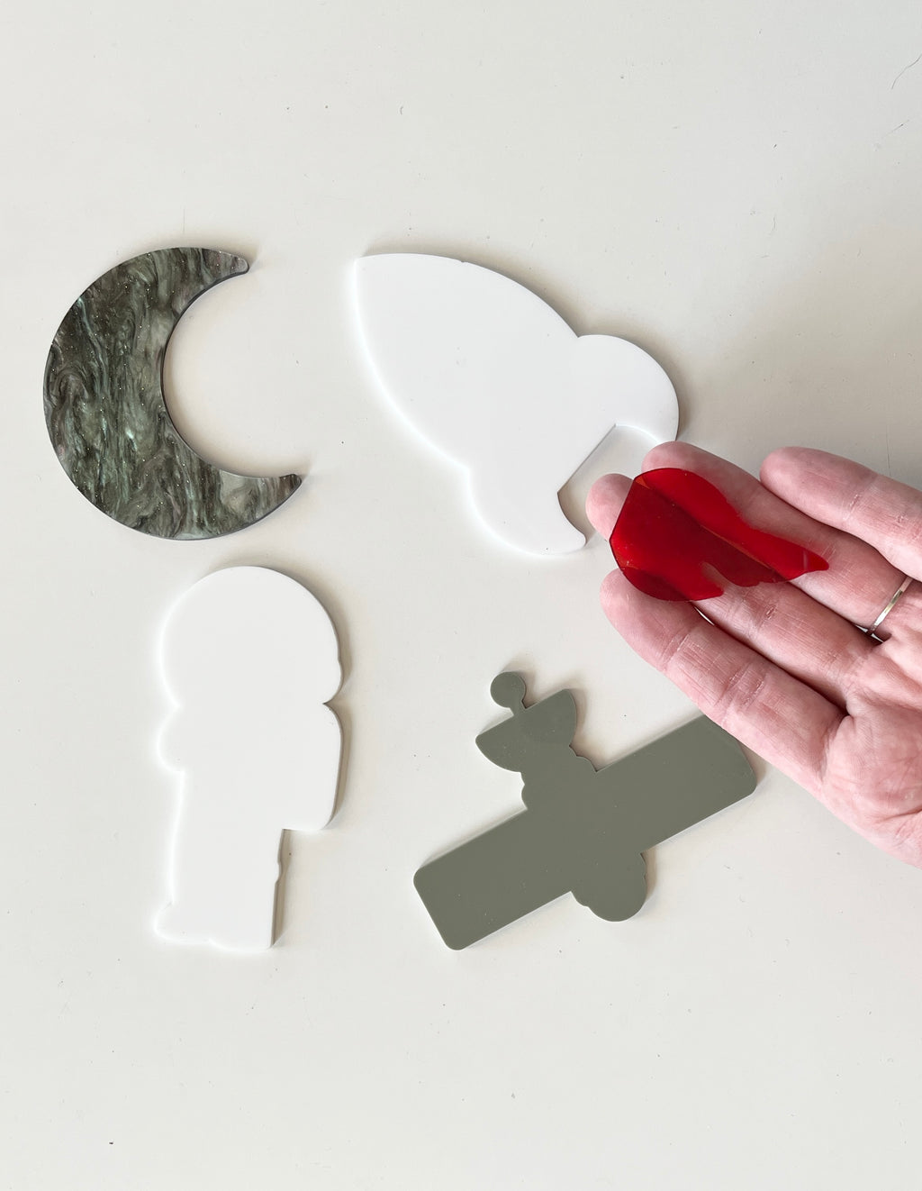 Hand holding red acrylic flame piece above white astronaut, gray satellite, white bird, and green marble crescent moon shapes on white surface