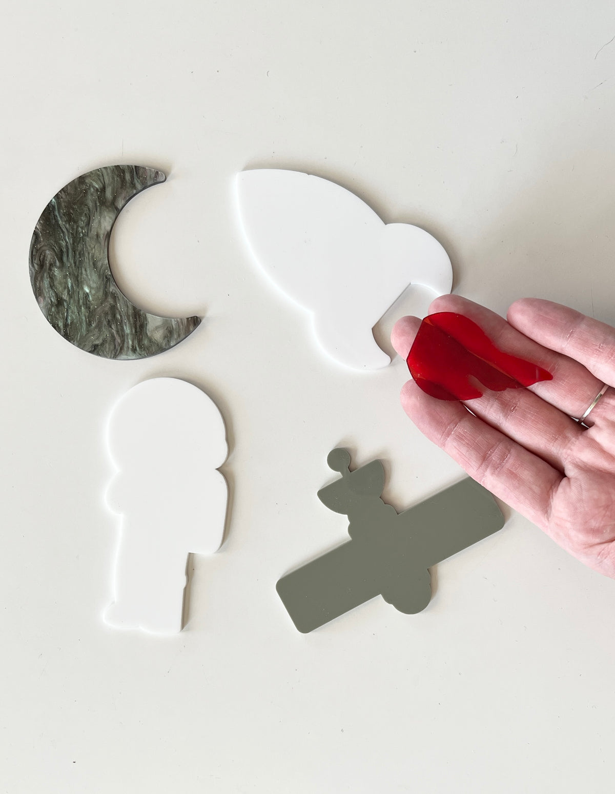 Hand holding red acrylic flame piece above white astronaut, gray satellite, white bird, and green marble crescent moon shapes on white surface