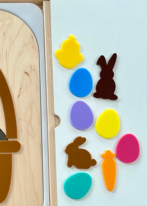 Colorful plastic Easter shapes including chicks, eggs, bunnies, and a carrot arranged on a white surface next to wood panel