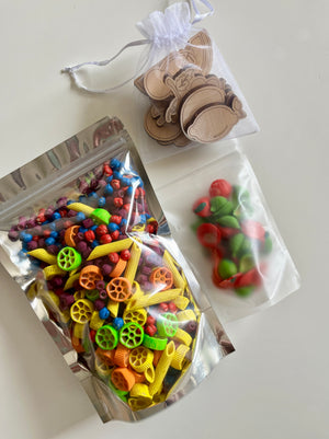 Clear resealable bags filled with colorful shaped pasta and a small white mesh pouch holding wooden cutouts on a light surface