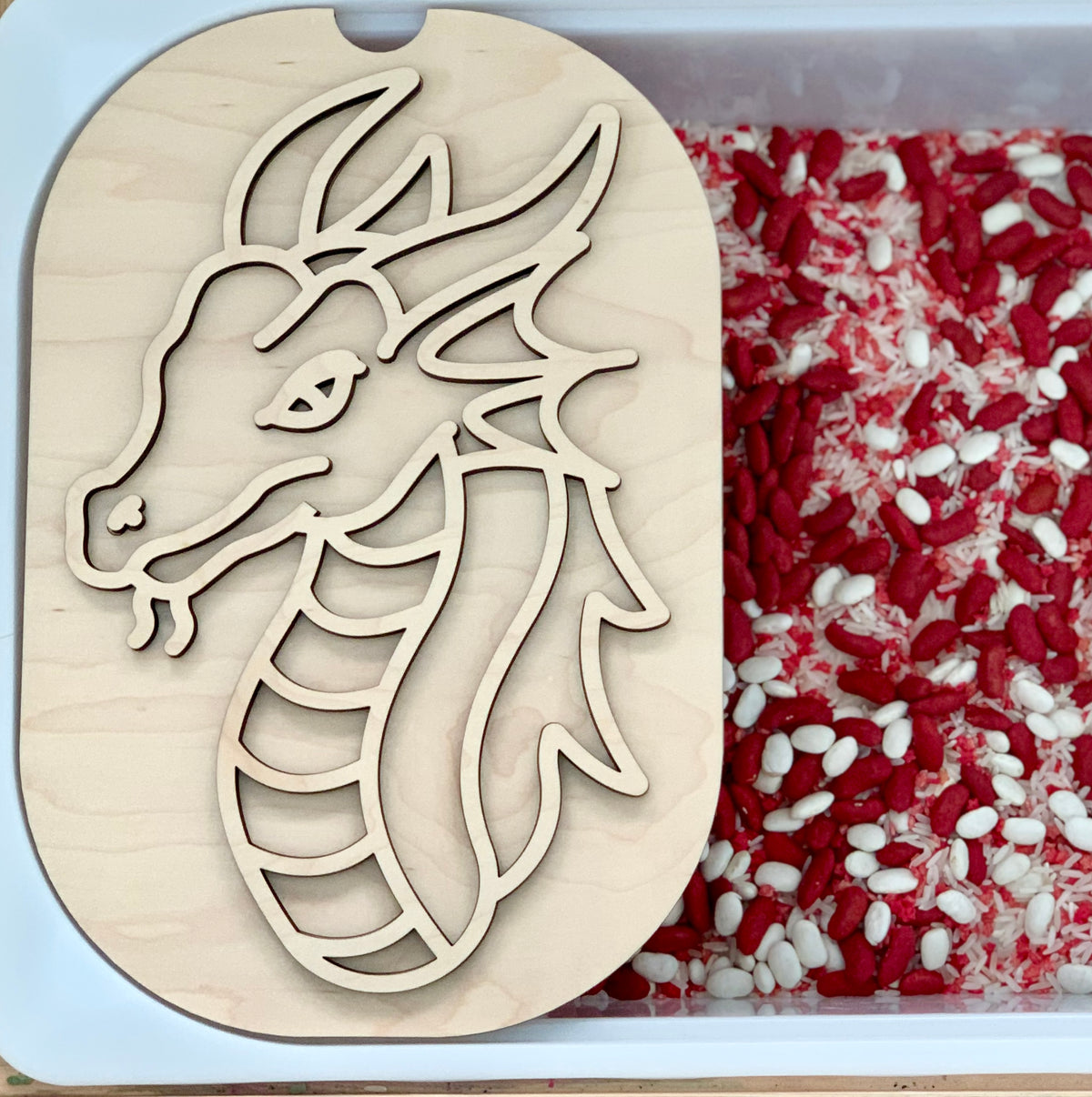 Wooden dragon stencil with carved outline resting on plastic container filled with red and white rice grains