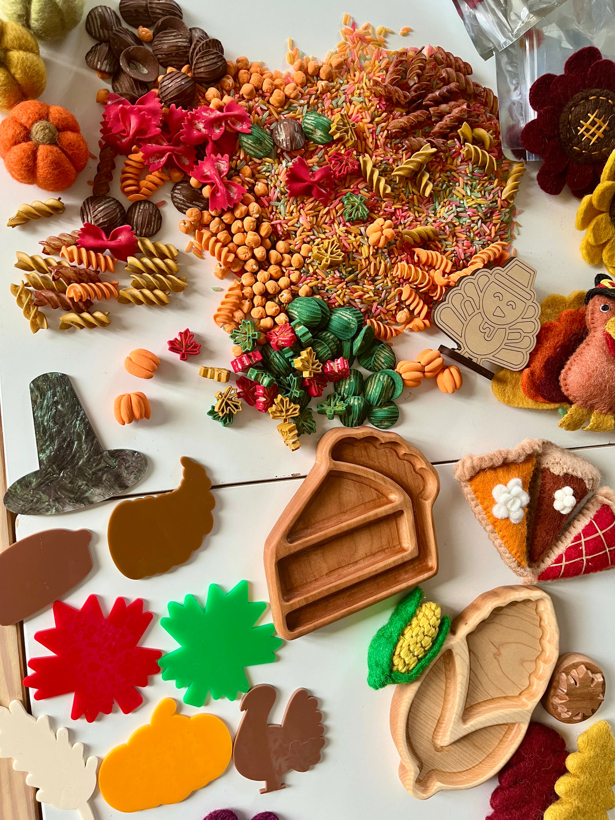 Assortment of autumn-themed craft supplies including colorful pasta, wooden trays, felt pumpkins, pie slices, and decorative leaves on white surface