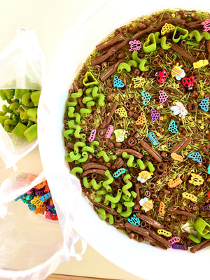 White bowl filled with multicolored insect-shaped cutouts and green and brown pasta pieces next to mesh bags