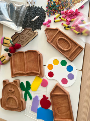Wooden trays in shapes of a book, schoolhouse, backpack, and pencil with colorful craft supplies on a white table