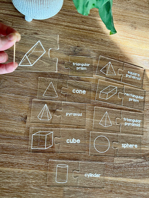Little Dry Erasables - 2 Part Geometry 3D Shape Puzzles display clear puzzle pieces with white illustrations and labels of 3D geometric shapes on a wooden table, handcrafted homeschool education resources.