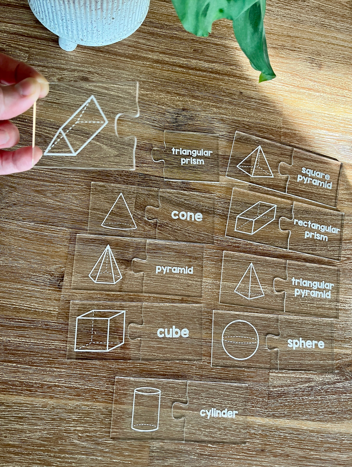 Little Dry Erasables - 2 Part Geometry 3D Shape Puzzles display clear puzzle pieces with white illustrations and labels of 3D geometric shapes on a wooden table, handcrafted homeschool education resources.