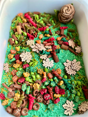 White bin filled with green rice, colorful pasta shapes, and wooden cutout leaves with a brown felt pinecone nearby
