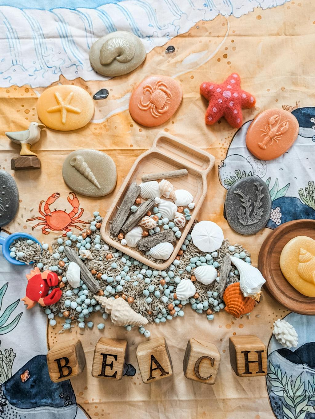 Natural wooden 1.5-inch alphabet blocks spelling BEACH surrounded by seashells, driftwood, and colorful beach-themed toys on fabric background