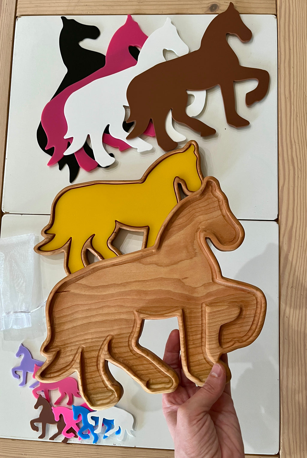 Stack of horse-shaped trays and flat cutouts in various colors including natural wood, brown, yellow, black, white, and pink on wooden table