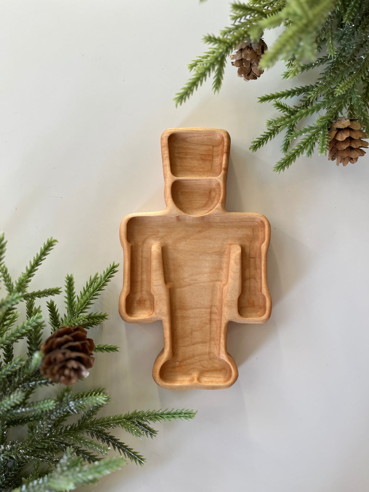 Light wooden tray carved in a robot shape surrounded by pine branches and pinecones on a white background