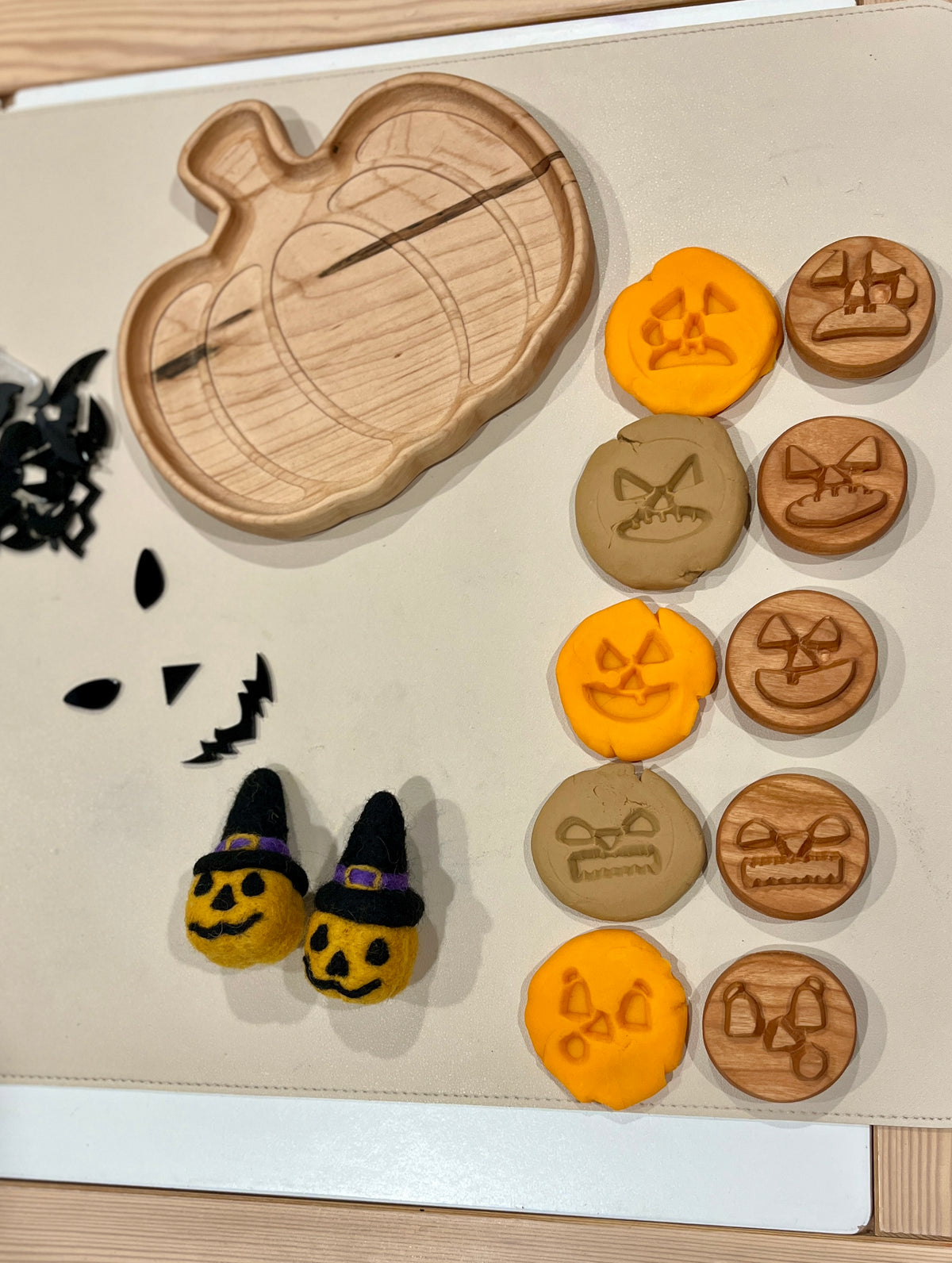 Wooden pumpkin-shaped tray with clay and wooden discs featuring various Halloween-themed carved faces and two felt pumpkin figures wearing witch hats