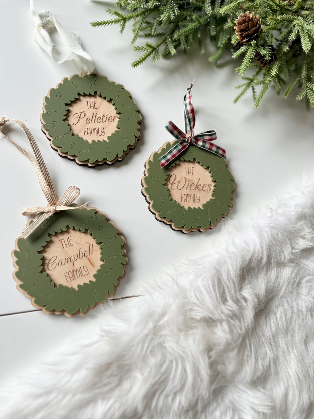 Three layered wooden wreath ornaments with green leaves and personalized family names on white surface next to greenery and fur rug
