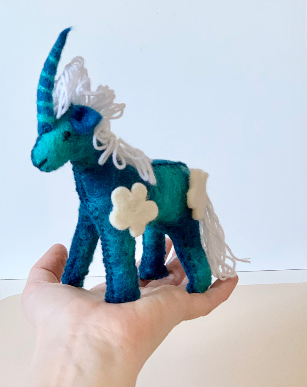 Hand holding a blue and teal felt unicorn with white yarn mane, tail, and cream felt flower patches against a plain background
