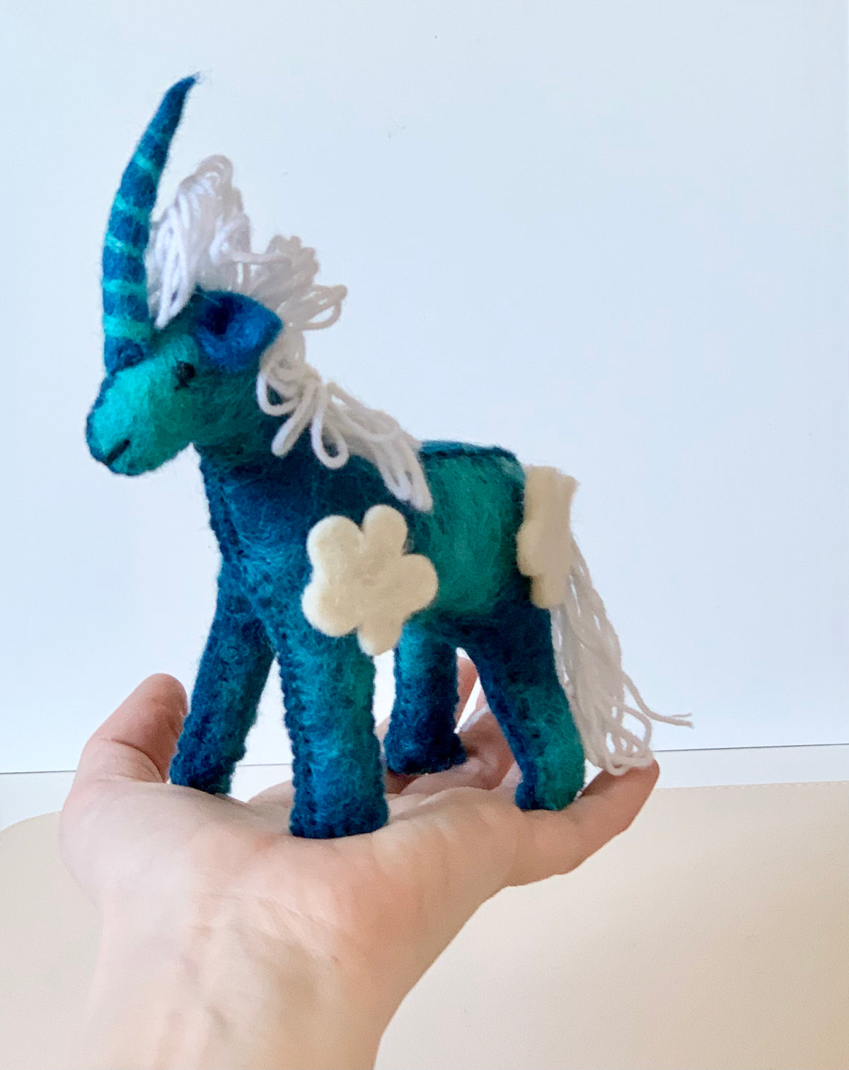 Hand holding a blue and teal felt unicorn with white yarn mane, tail, and cream felt flower patches against a plain background