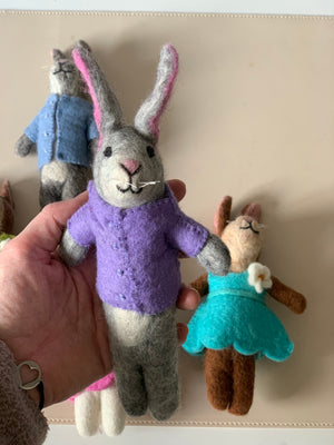 Handcrafted gray felt bunny doll wearing a purple outfit held in hand with two other felt animal dolls in the background
