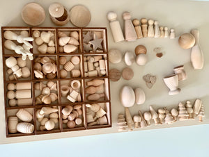 Unpainted wooden toys and shapes including peg dolls, animals, eggs, stars, and trees arranged on a beige surface and in a tray