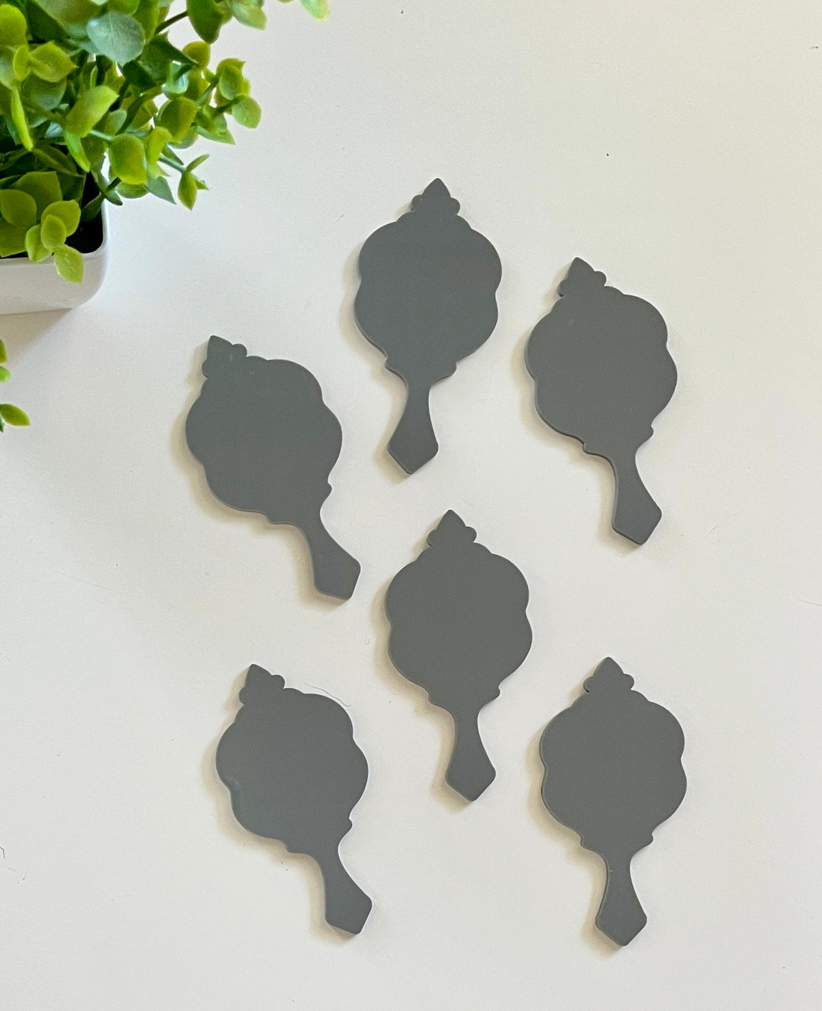 Six small black decorative shapes with ornate tops arranged on a white surface next to a green plant