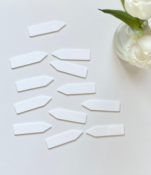 Scattered white plastic plant labels on a white surface next to a glass vase holding white flowers and green leaves