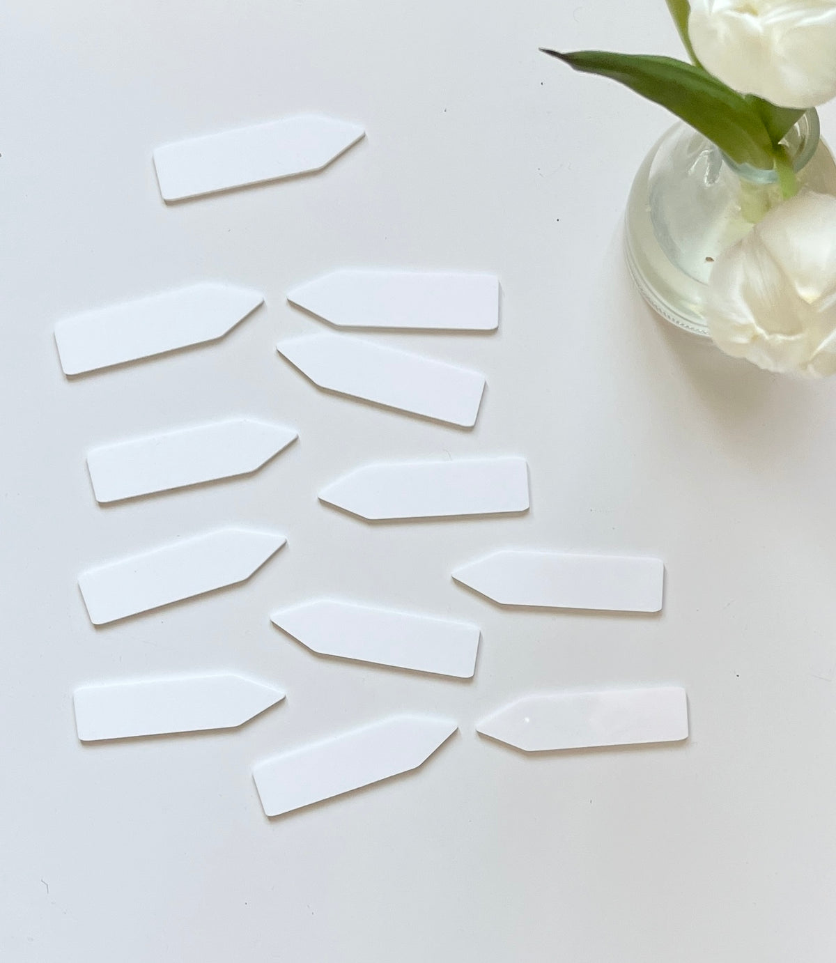Scattered white plastic plant labels on a white surface next to a glass vase holding white flowers and green leaves