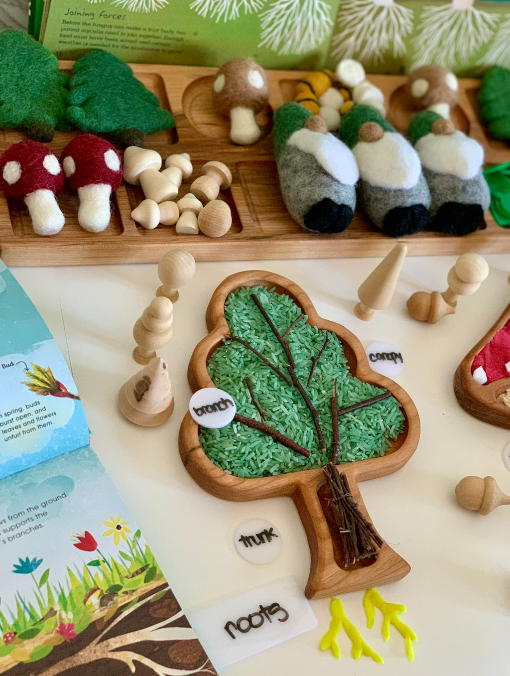Tree-shaped wooden sensory tray filled with green rice, small wooden figures, felt mushrooms, and gnome toys on white table
