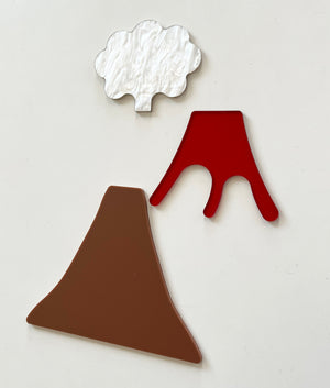 Three flat acrylic shapes including a brown volcano base, red lava flow with branches, and textured white smoke cloud on light background