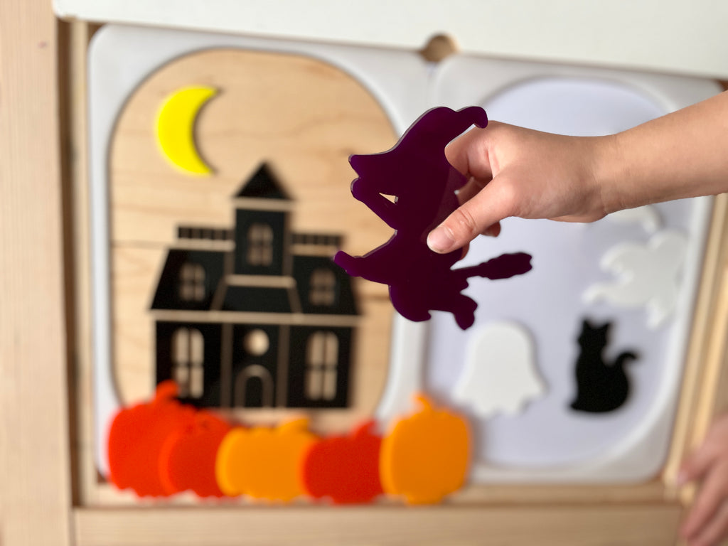 Child's hand holding a purple witch piece above a wooden puzzle with a haunted house, crescent moon, and pumpkins on a light wood frame