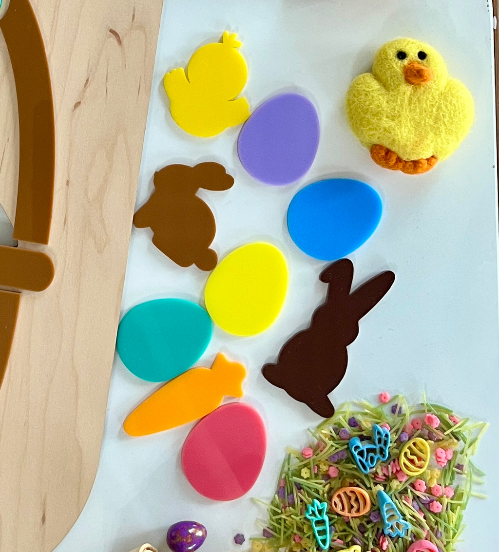 Assorted colorful plastic Easter shapes including eggs, bunnies, a chick, and a carrot on a white surface next to a wooden tray