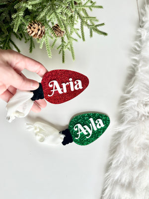 Hand holding red glittery Christmas light-shaped ornament with white name near green glittery light-shaped ornament on white surface