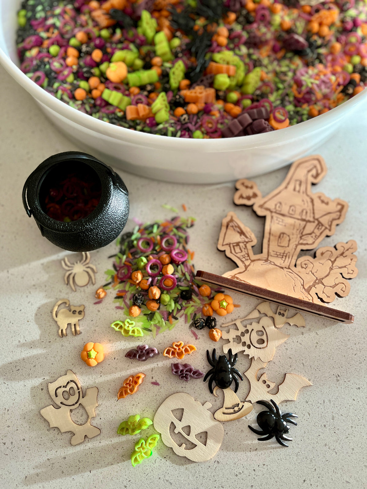 White bowl filled with colorful Halloween pasta shapes and wooden spooky house with assorted Halloween-themed decorations nearby