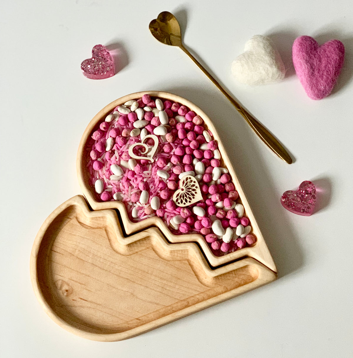 Broken heart-shaped wooden tray filled with pink and white sensory beads, felt hearts, and a gold heart-topped spoon nearby