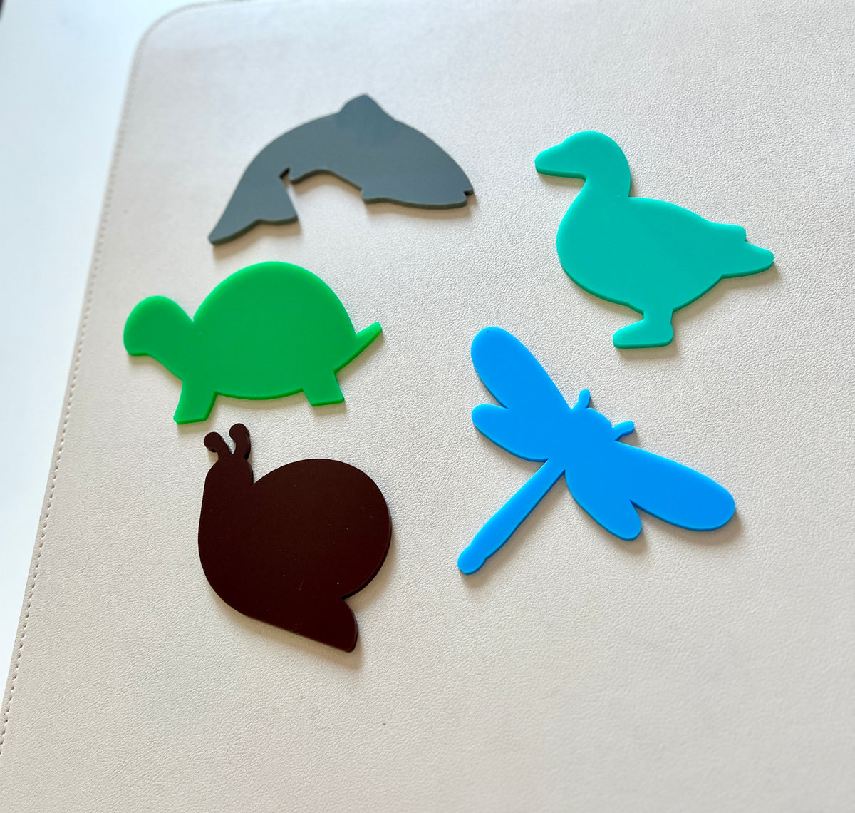 Colorful animal-shaped silicone figures including a dolphin, duck, turtle, dragonfly, and snail on a cream surface