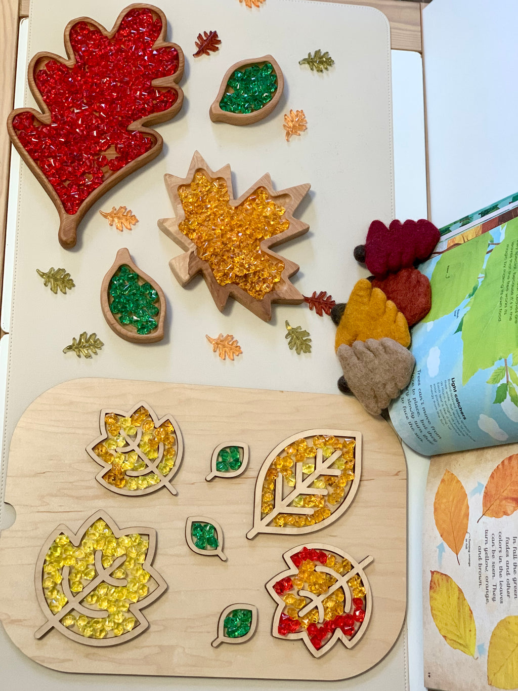 Wooden leaf-shaped sensory trays filled with colorful beads, felt leaves, and an open book on white surface