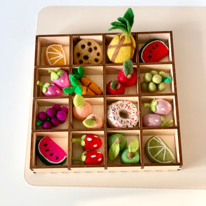 Wooden grid with compartments holding colorful felt fruits, vegetables, and a donut with sprinkles on a white background