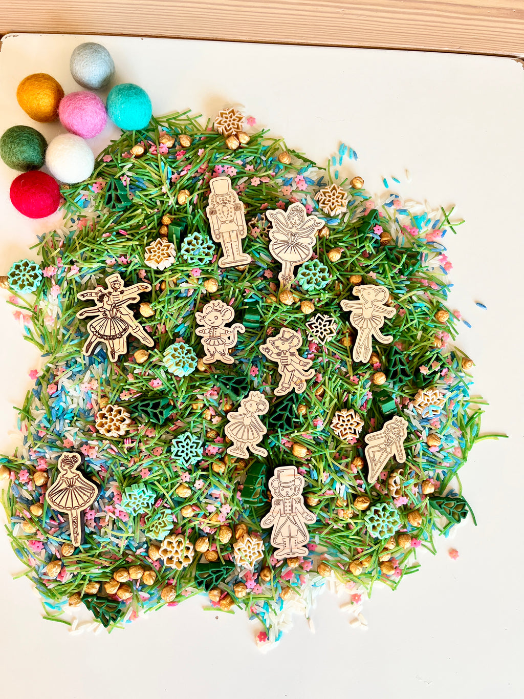 Wooden Nutcracker and ballerina cutouts on green pine needle mix with colorful sprinkles and small gold beads on white surface
