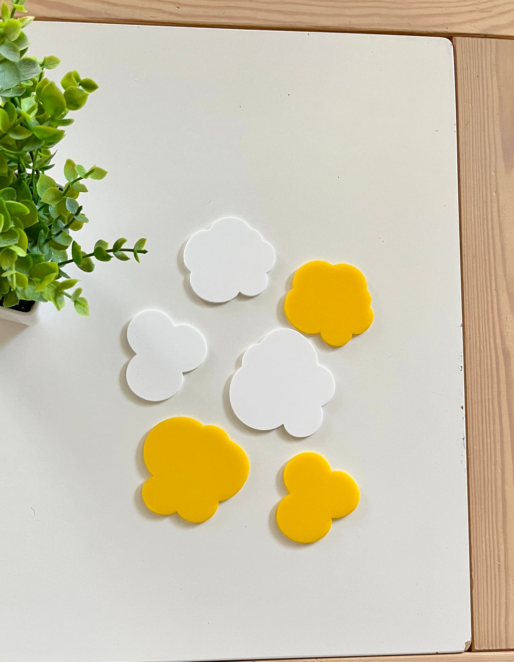 Seven cloud-shaped cutouts in white and yellow arranged on a white surface near a small green plant