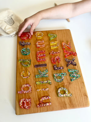 Child’s hand reaching for colorful resin uppercase alphabet letters arranged on a wooden board with a fabric pouch nearby