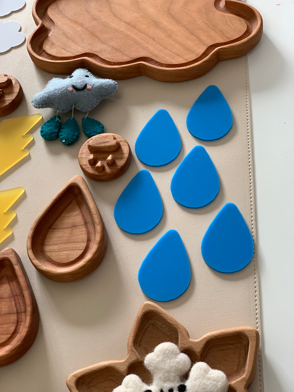 Six blue raindrop-shaped pieces arranged on a beige surface near wooden trays and a small plush cloud toy