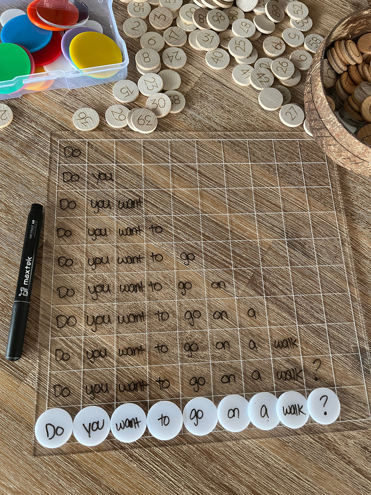 Dry Erasable Blank Open Ended 100 Frame Hundred Board on wood surface with black marker, circular number tokens, colorful translucent discs, and white letter discs spelling a question, handcrafted homeschool education resources.