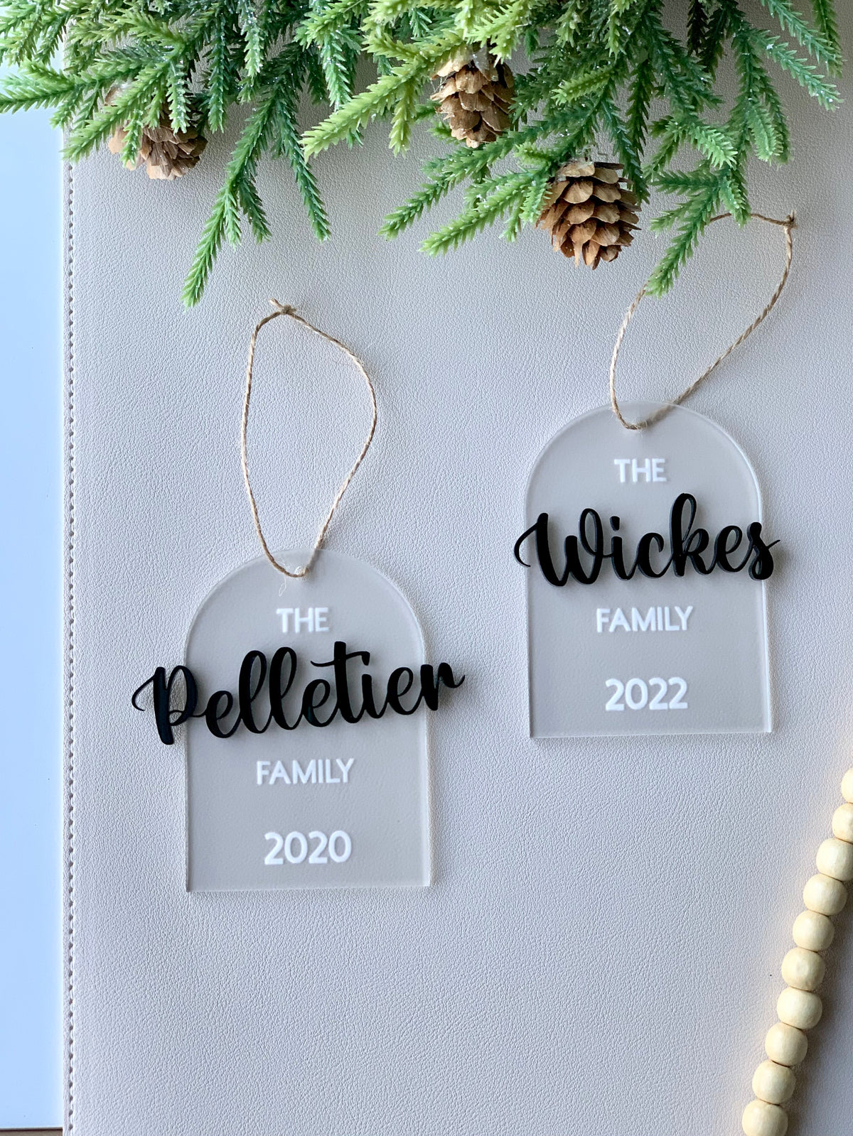 Two clear acrylic arch ornaments with black cursive family names and white text showing years, hanging from twine below pine branches