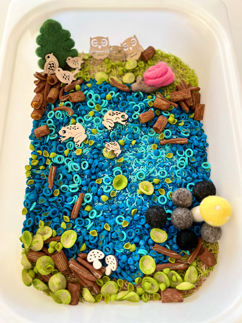Sensory bin with blue pasta “pond,” wooden frogs and birds, felt tree and snail, green moss and sticks scattered around.